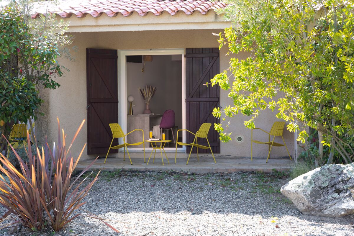 A covered outdoor seating area with yellow chairs and a table, a rock, and a plant.