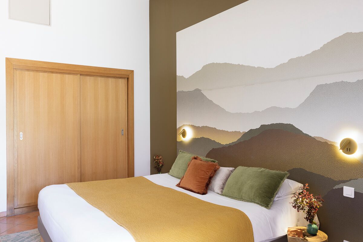 Un dormitorio con cama, almohadas, manta, una lámpara fijada a la pared y una puerta.