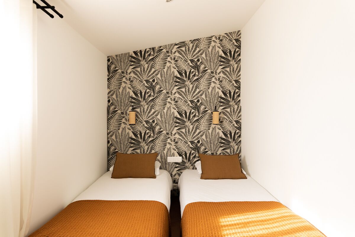 A small, narrow room with two single beds, a patterned wallpaper, and a simple, clean design.