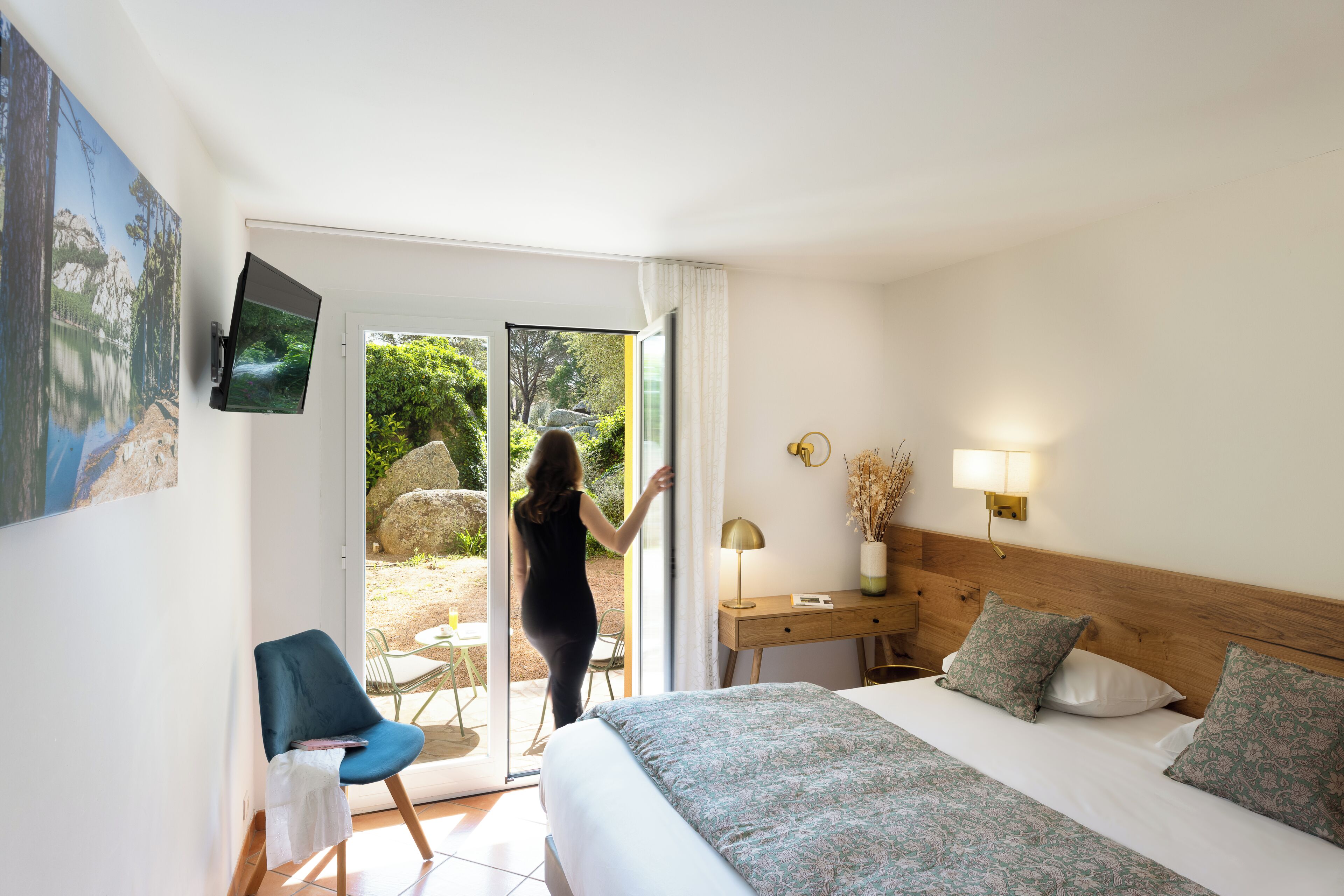 Habitación de hotel con cama, escritorio, silla, televisión y puerta corrediza de vidrio que da a un espacio al aire libre.
