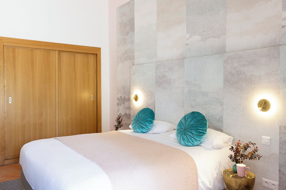Une chambre avec une armoire en bois, un lit avec une literie blanche, des coussins turquoise et des lampes fixées au mur.