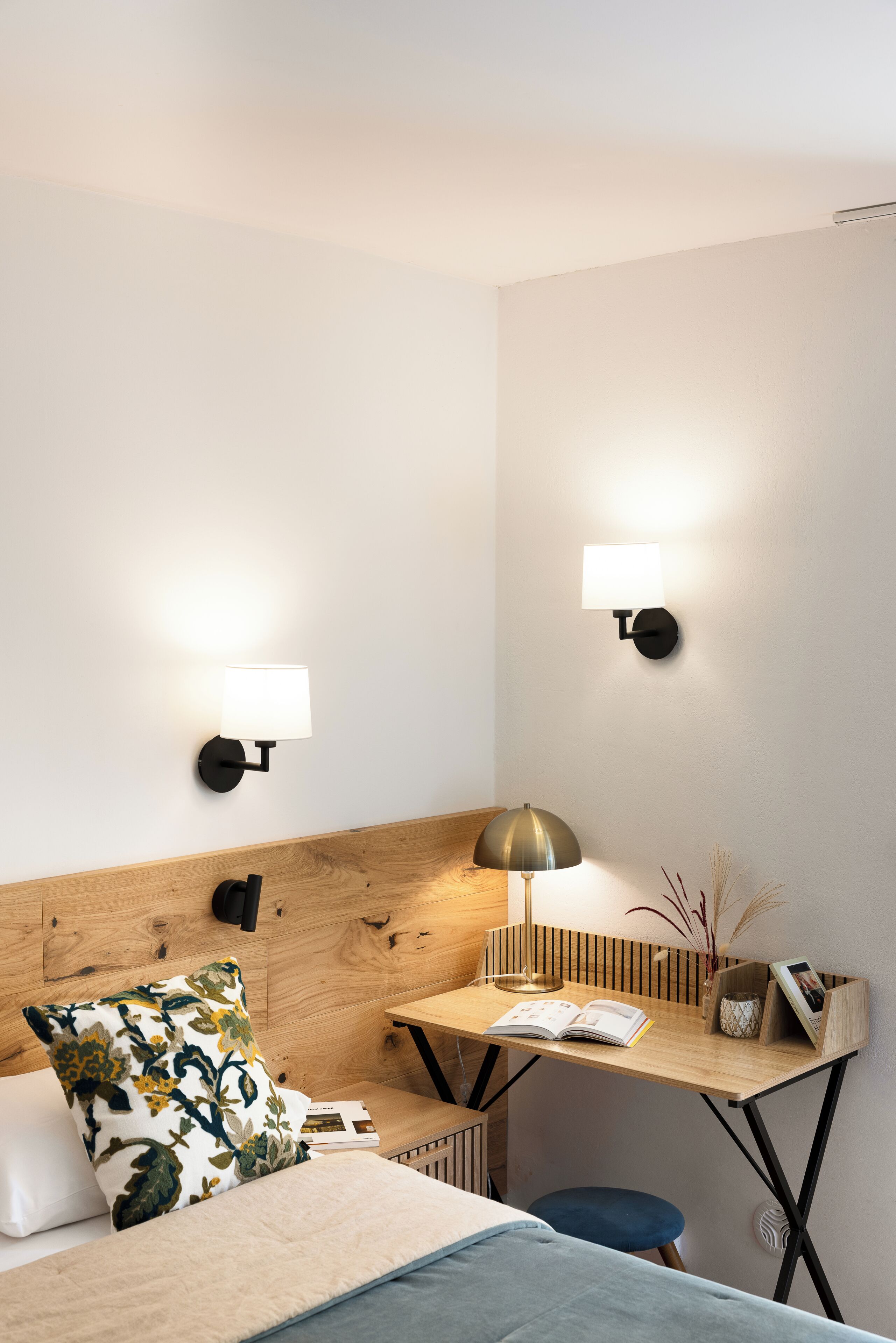 A hotel room with a bed, a desk with a lamp, a chair, a small table with books and a vase, and two wall-mounted lights.