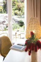 Una habitación con un ventanal amplio, una lámpara de mimbre, un sillón amarillo y un jarrón con flores rojas.