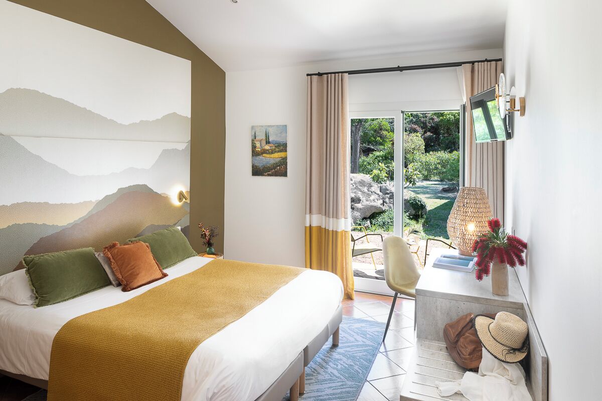 Une chambre d’hôtel avec un lit, un bureau avec une lampe, une télévision et une vue sur un jardin.