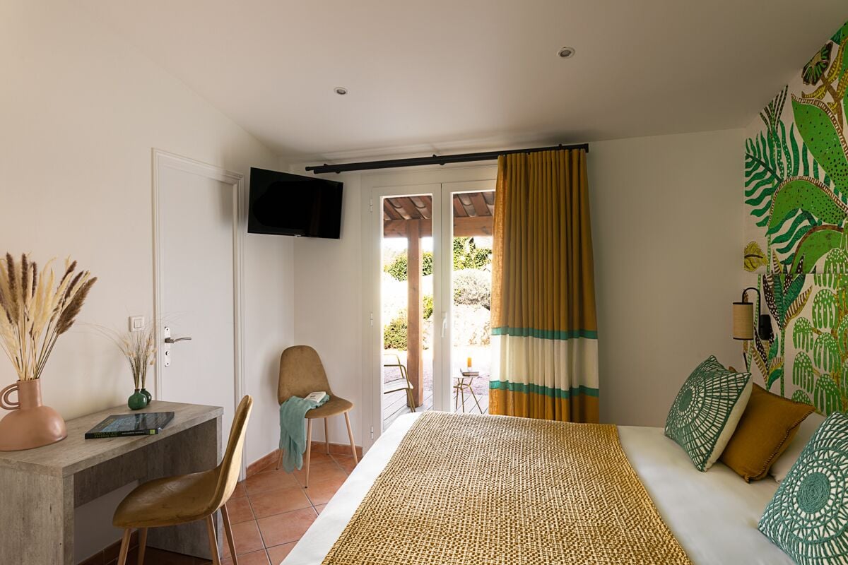 Quarto com cama, escrivaninha, cadeira, televisão e vista para o exterior.