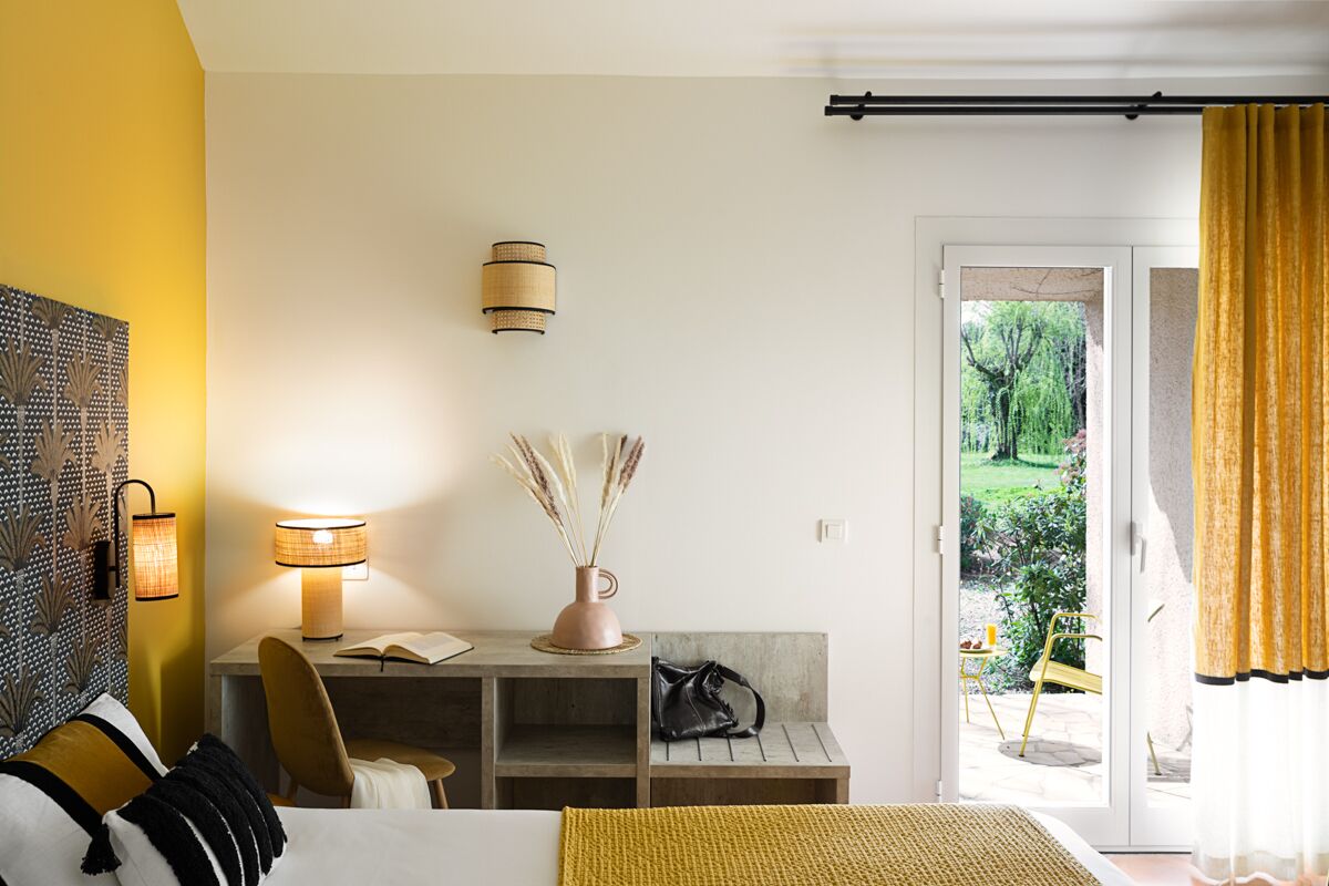 A hotel room with a bed, a desk with a lamp, a vase with pampas grass, and a view of a garden through a sliding door.
