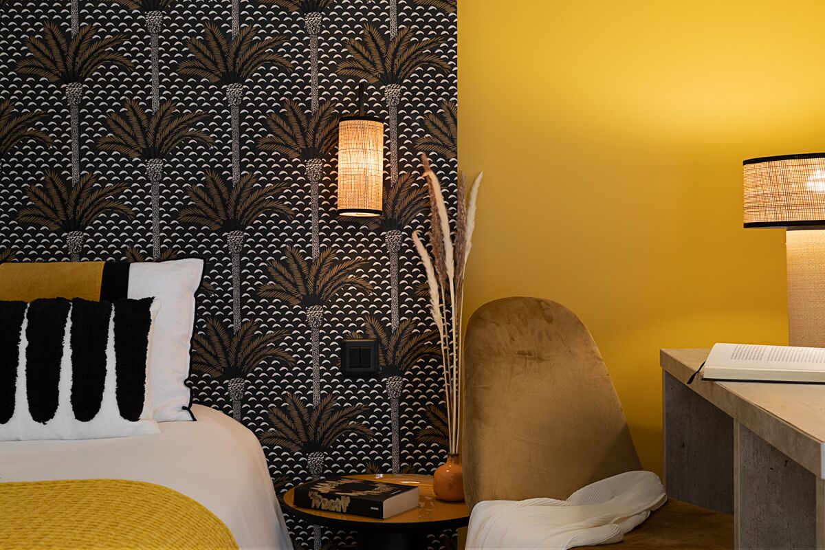 A hotel room with a patterned wallpaper, a bed with black and white pillows, a round side table with a book and a lamp, and a wooden chair.