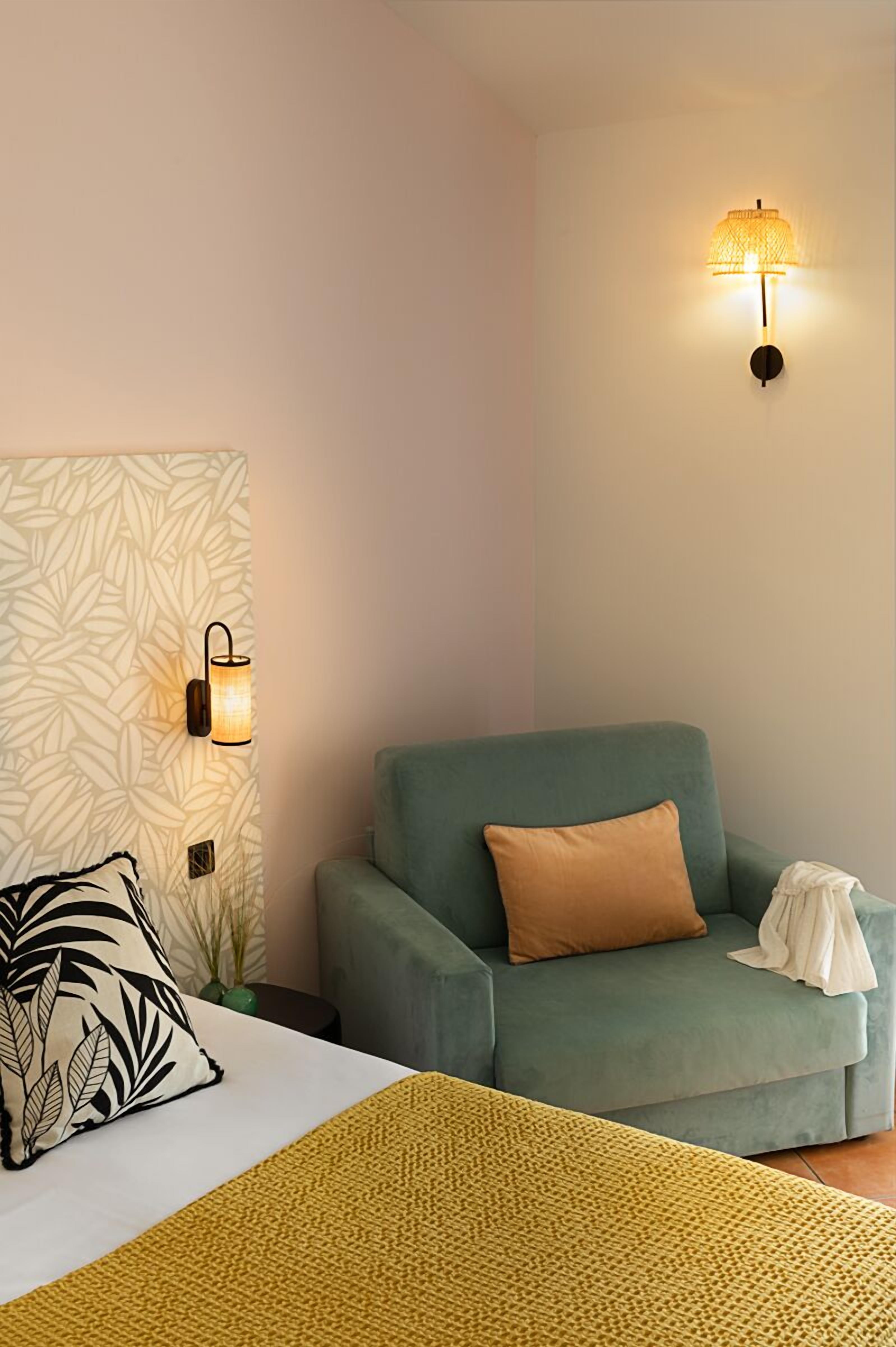 Um quarto com uma cama, uma poltrona verde, parede estampada e duas luminárias fixadas na parede.