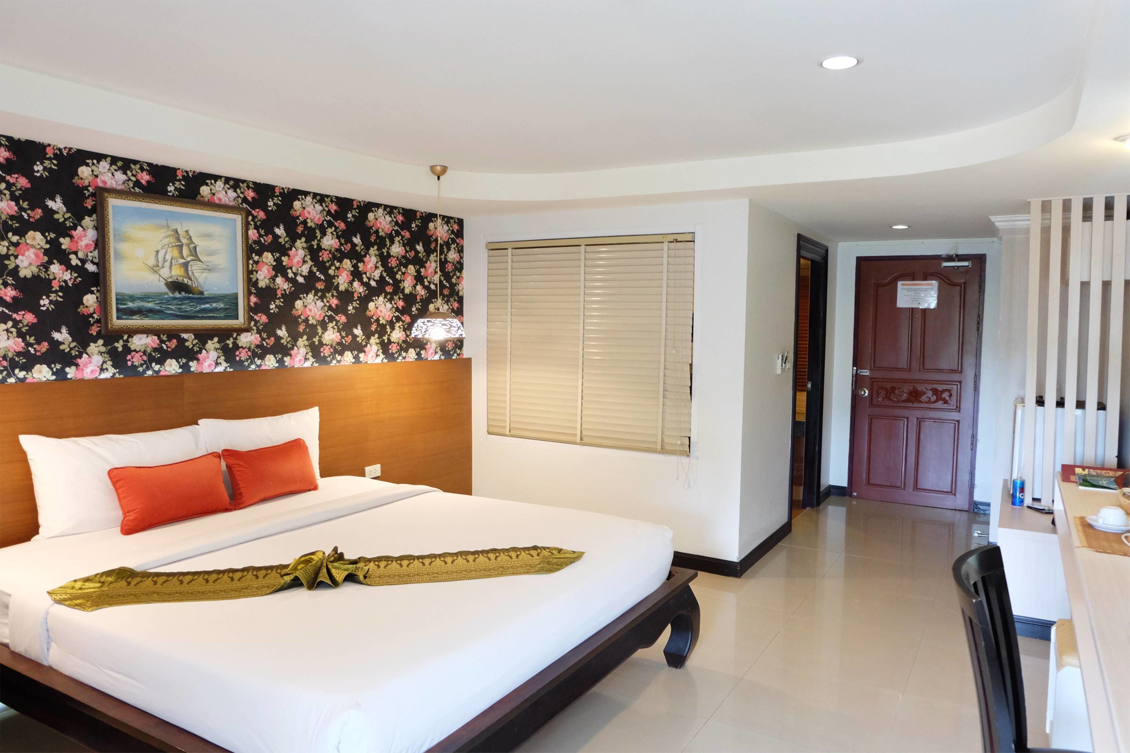 A hotel room with a large bed, a wooden headboard, a floral wallpaper, a painting, a window with blinds, and a door.