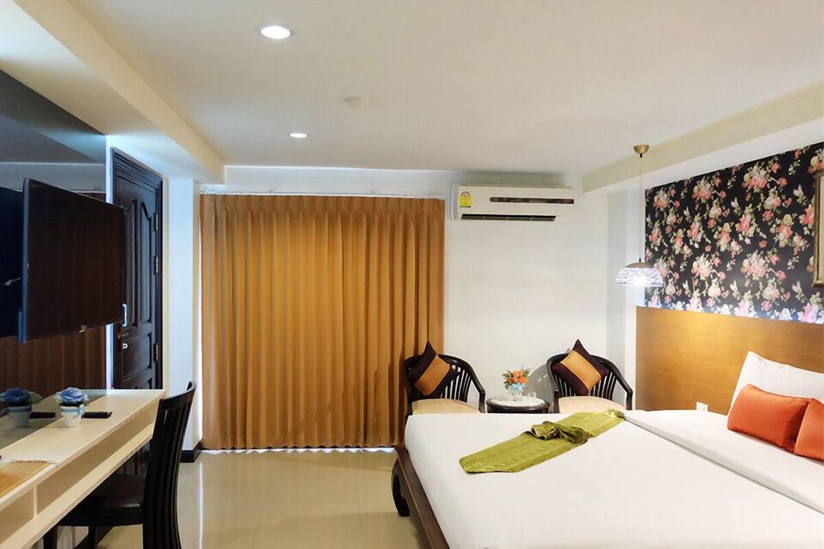 A hotel room with a bed, a desk, two chairs, a TV, and an air conditioning unit.