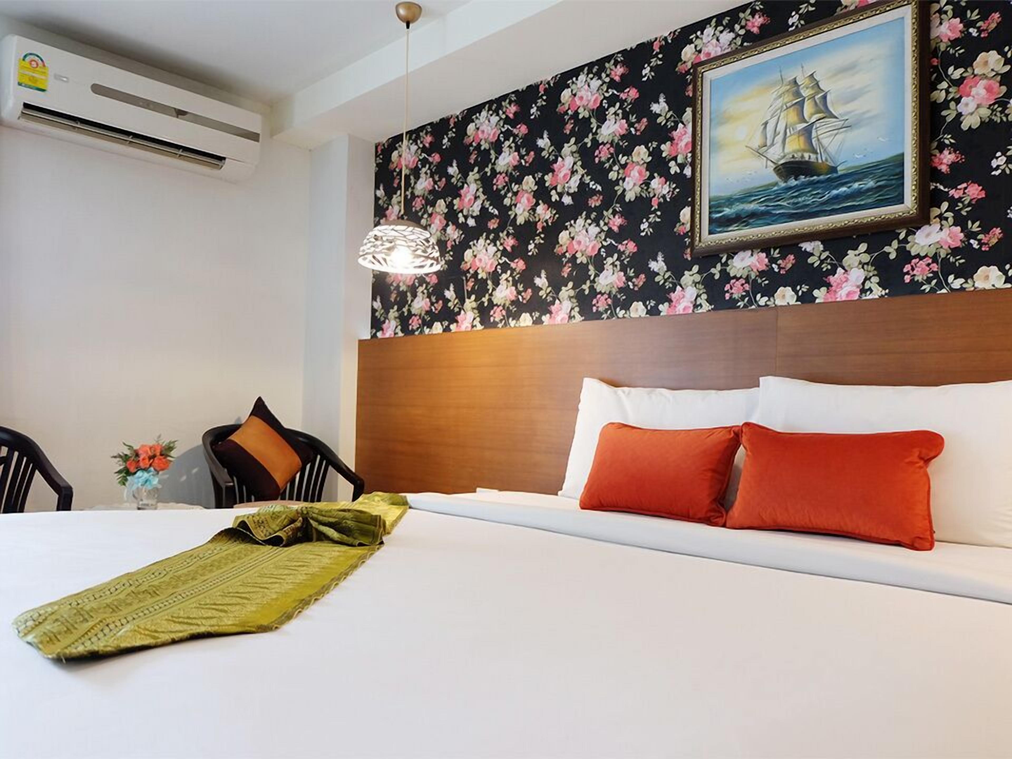A hotel room with a bed, a floral wallpaper, a painting of a ship, and a ceiling fan.
