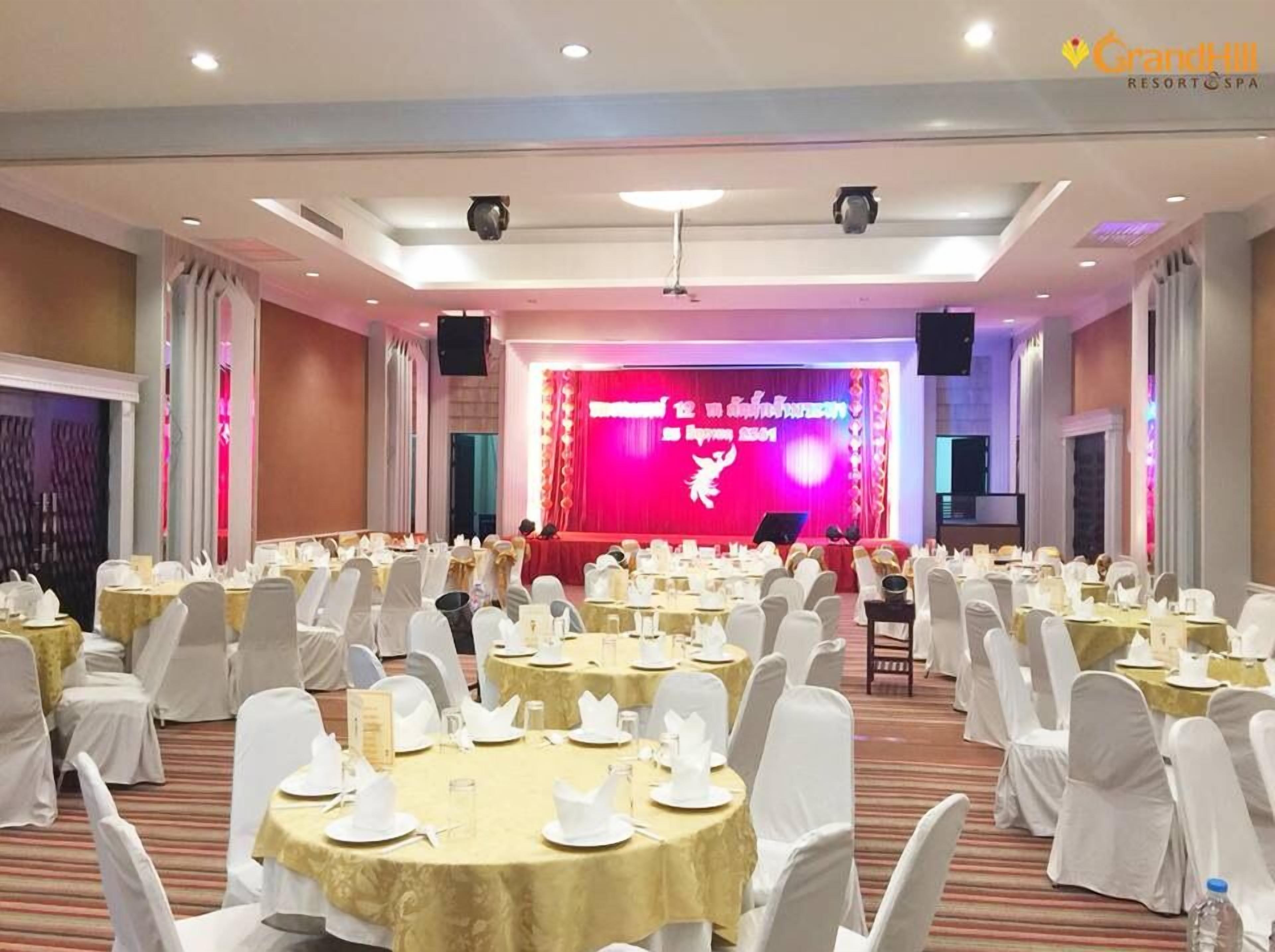 Un salón de banquetes con mesas redondas preparadas para un evento formal, un escenario con un telón rojo, y el logotipo de GrandHill Resort & Spa.