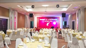 Un salón de banquetes con mesas redondas preparadas para un evento formal, un escenario con un telón rojo, y el logotipo de GrandHill Resort & Spa.