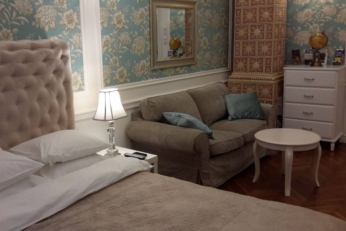 A bedroom with a tufted headboard, a bed with white linens, a nightstand with a lamp, a brown sofa with cushions, a small round table, and a dresser with drawers.