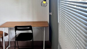 A small room with a wooden table and a black chair, a wall-mounted lamp, and a window with blinds.
