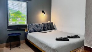 A bedroom with a bed, a window with blinds, an air conditioner, a wall-mounted light, and a small stool.