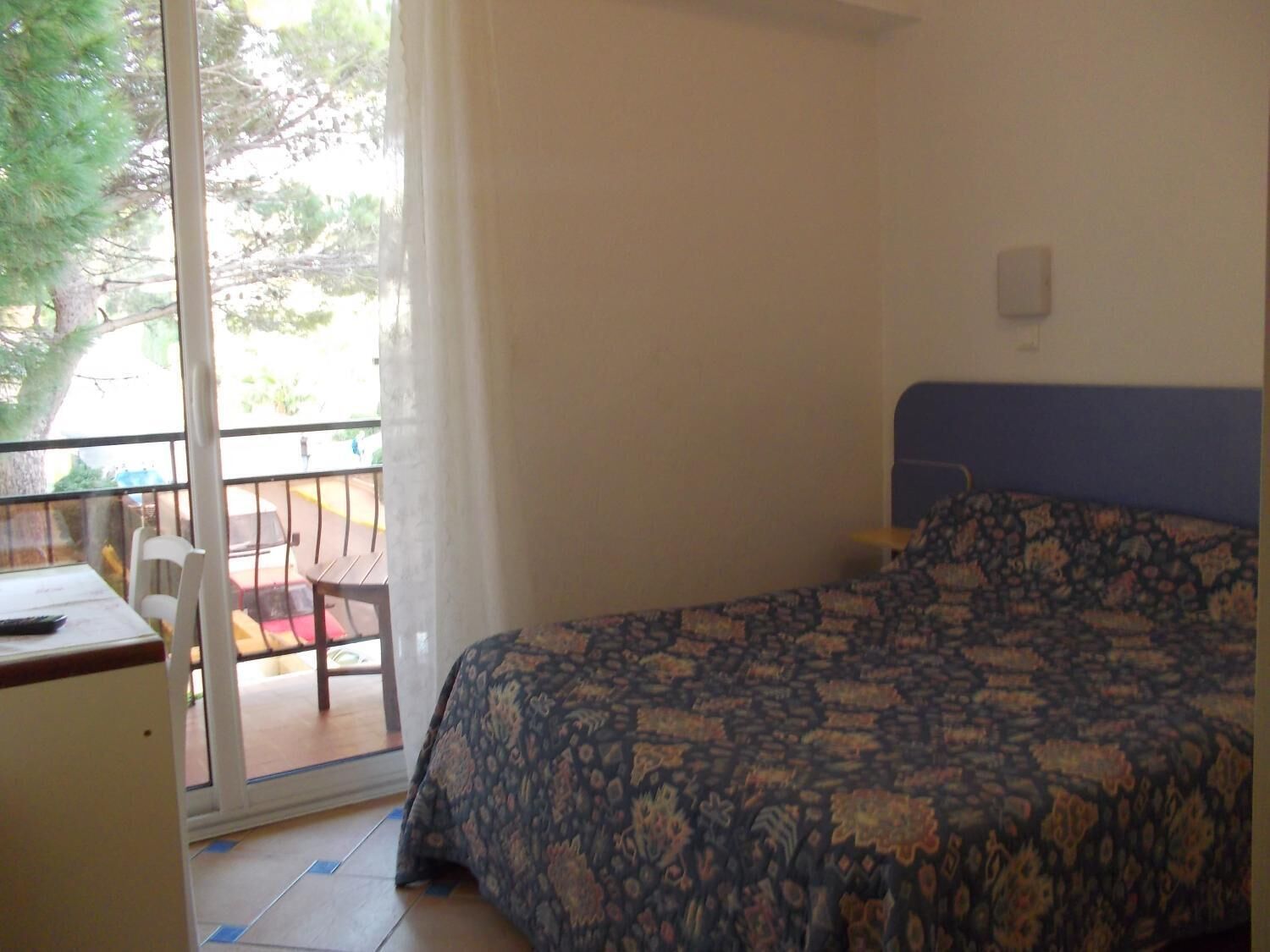 A bedroom with a bed, a desk, and a view of a balcony with chairs.