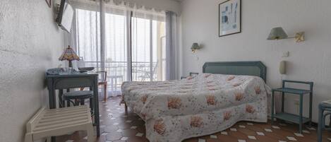 Un dormitorio con cama, escritorio, silla, mesita y balcón con vistas.