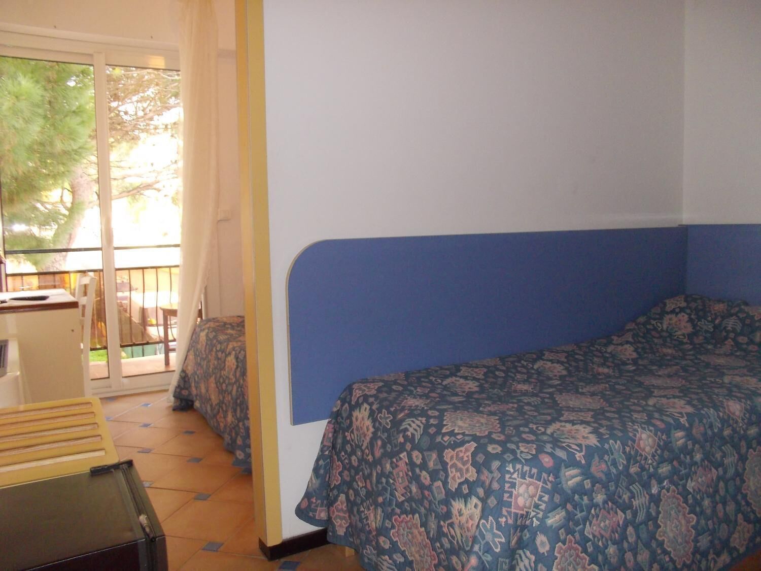 Un dormitorio con cama, un escritorio y una silla. Hay un balcón con vista a árboles.