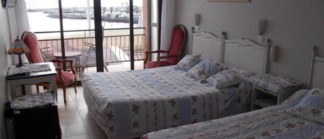 A hotel room with two beds, a desk, a chair, and a view of the sea through large windows.