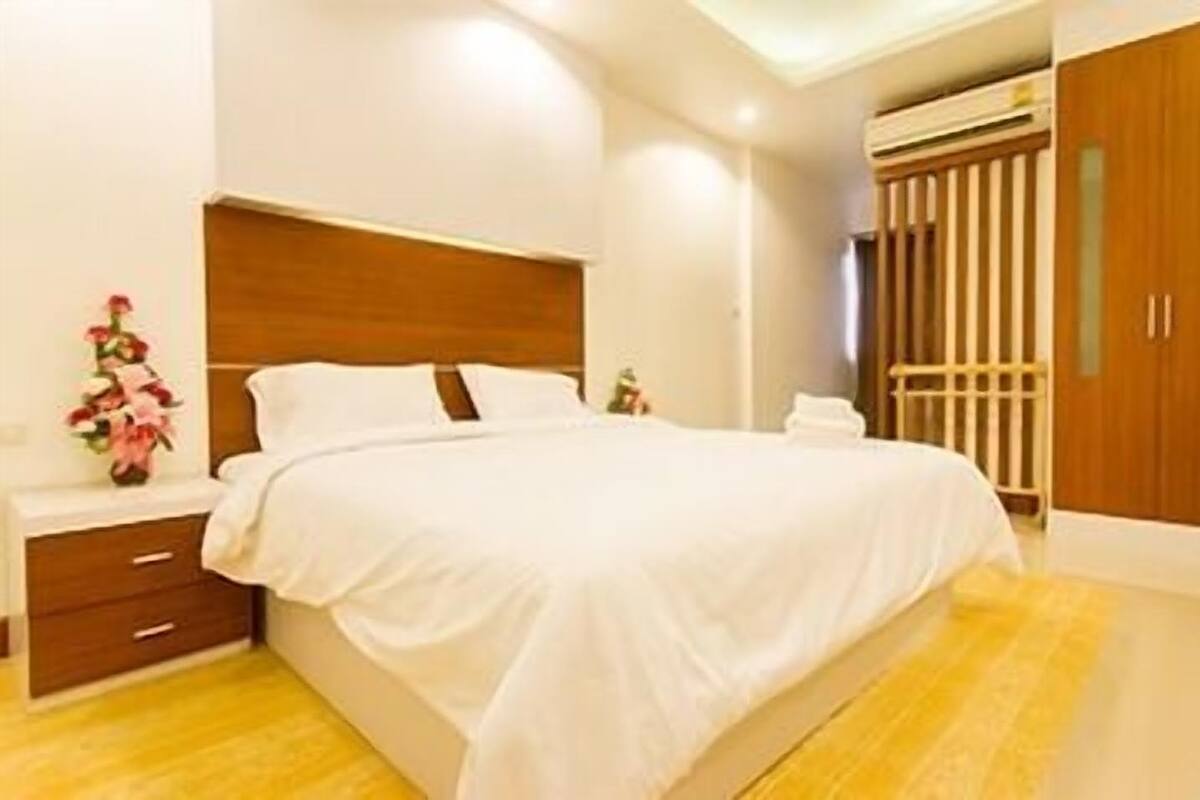 Ein Hotelzimmer mit einem großen Bett, einem Holz-Kopfschild, einem Nachttisch mit Schublade, einem kleinen Tisch mit Blumenvase und einem Kleiderschrank mit vertikalen Lamellen.