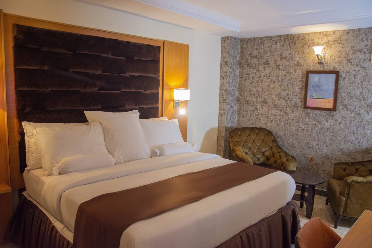 Hotellihuone, jossa on suuri sänky, pääty, yöpöytä lampun kanssa, pieni pöytä, kaksi nojatuolia ja seinällä oleva kehystetty kuva.