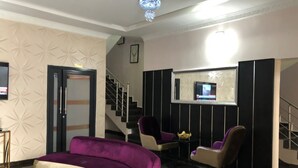 Eine Hotelhalle mit einer Rezeption, einem violetten Sofa, zwei Sesseln, einer Treppe und einem an der Wand befestigten Fernseher.
