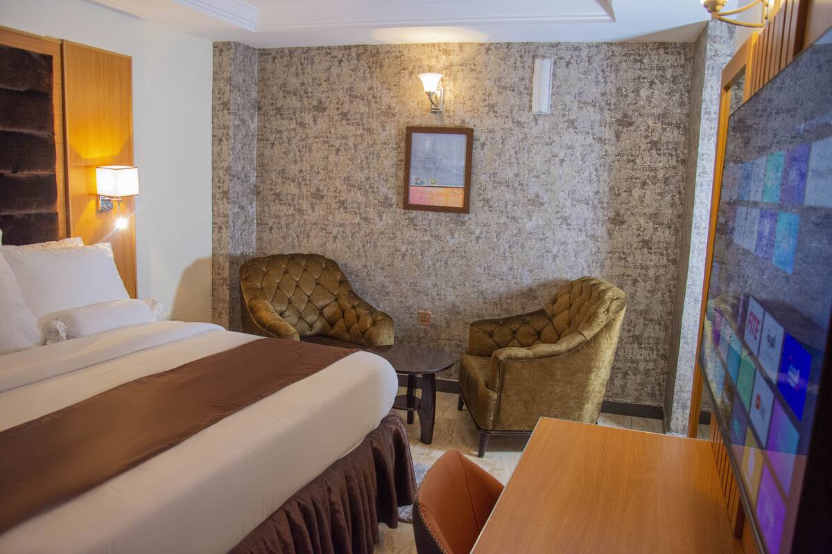 Hotellihuone, jossa on suuri sänky, kaksi nojatuolia, pieni pöytä ja seinään kiinnitetty lamppu.