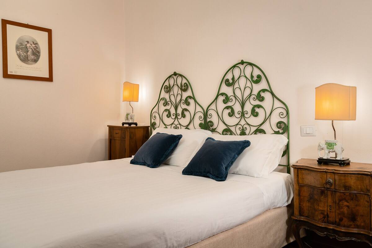 A neatly made bed with white linens and two dark blue pillows, a green metal headboard, a wooden nightstand with a lamp, and a framed picture on the wall.