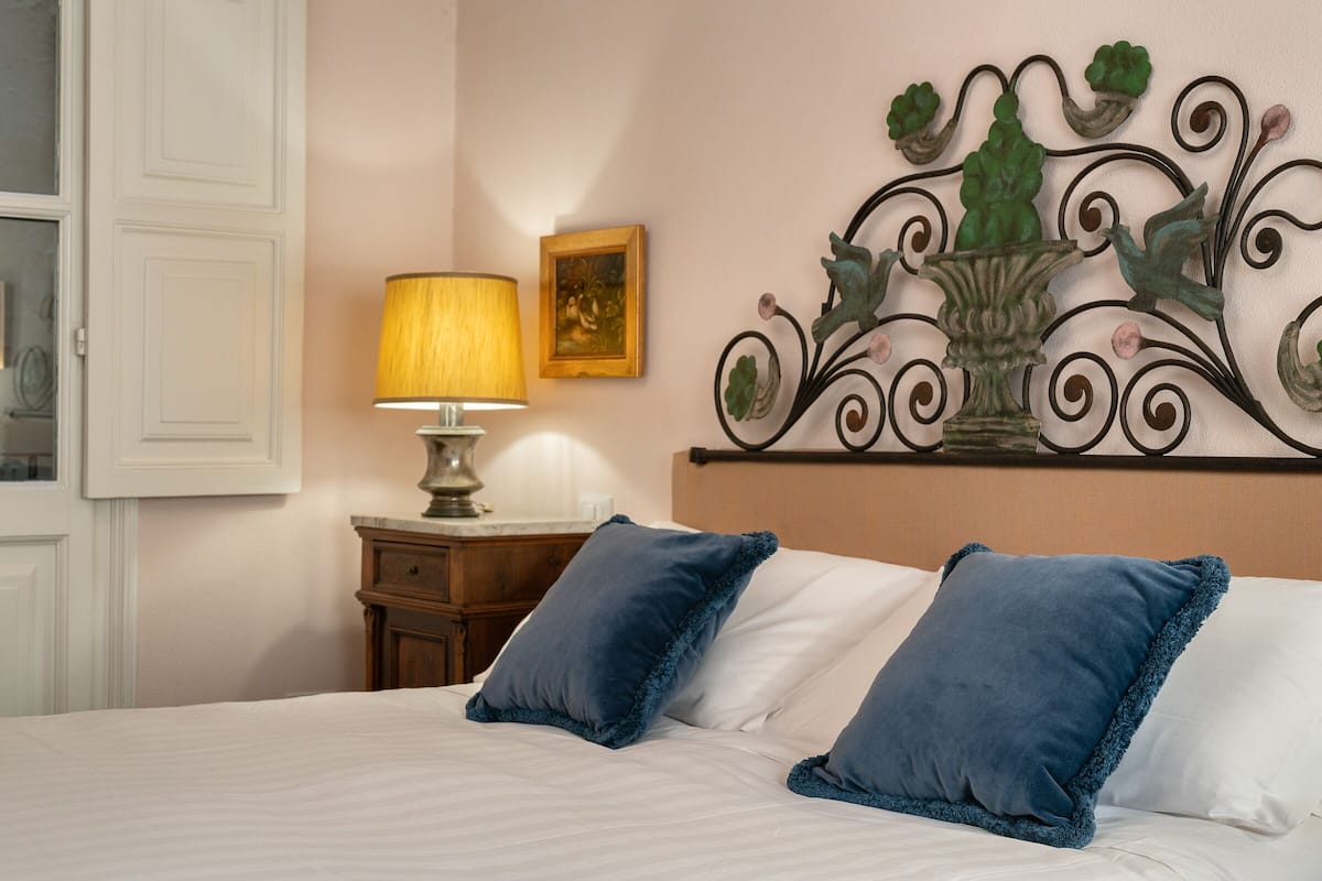 A neatly made bed with white and blue pillows, a bedside lamp, a wooden nightstand, and a decorative metal headboard with a floral design.
