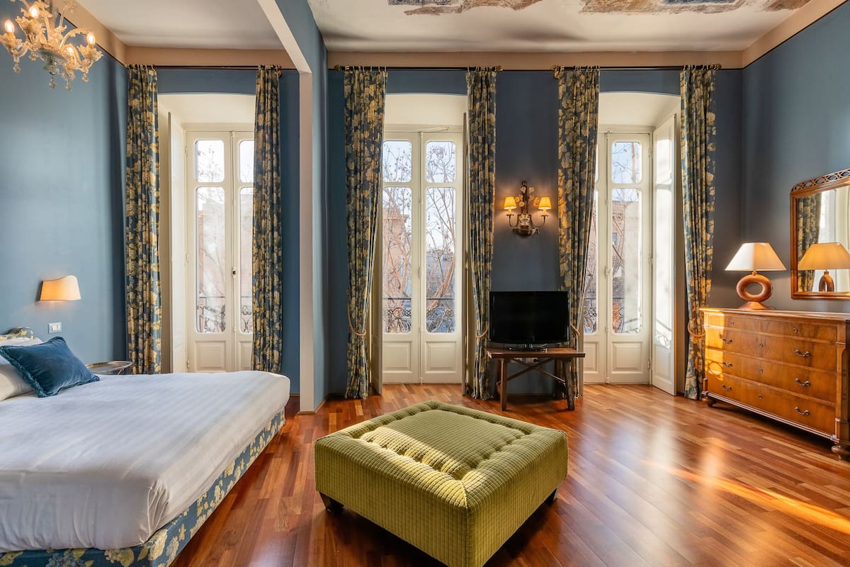 A bedroom with a bed, a green ottoman, a dresser, a TV, and a chandelier.