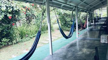 Un espacio al aire libre techado con hamacas, piso de concreto y un jardín con diversas plantas y flores.