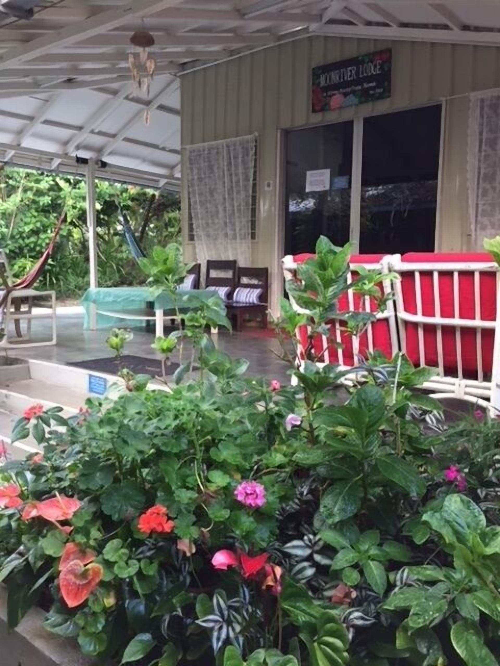 Un espace extérieur abrité, agrémenté d’une enseigne indiquant « MONTROSE LODGE ». On y trouve des coins salon avec des coussins rouges et blancs, ainsi qu’une variété de plantes et de fleurs en pot.