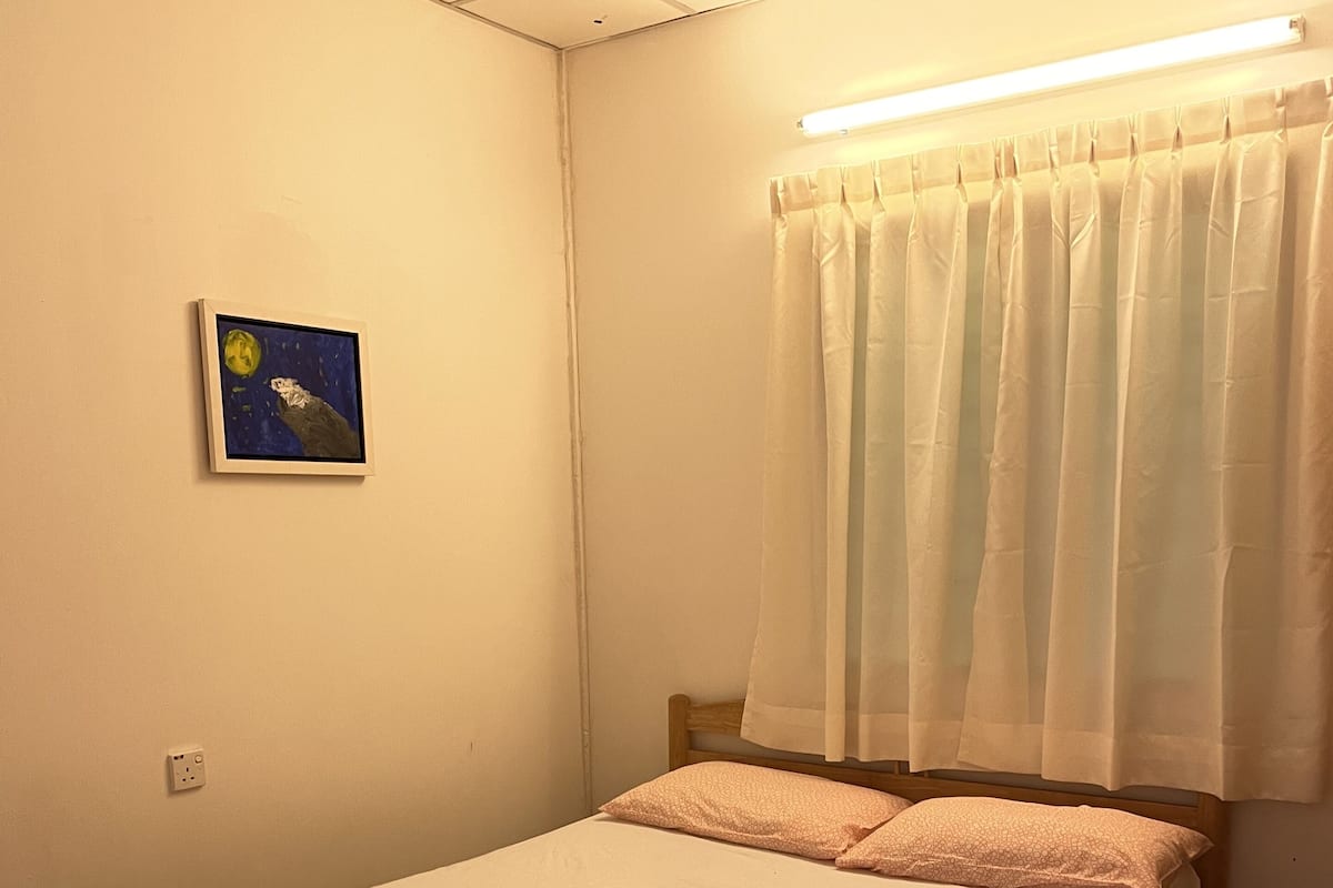 A single-bed room with a wooden frame, a white mattress, peach-colored pillows, a framed picture on the wall, and a window with sheer curtains.