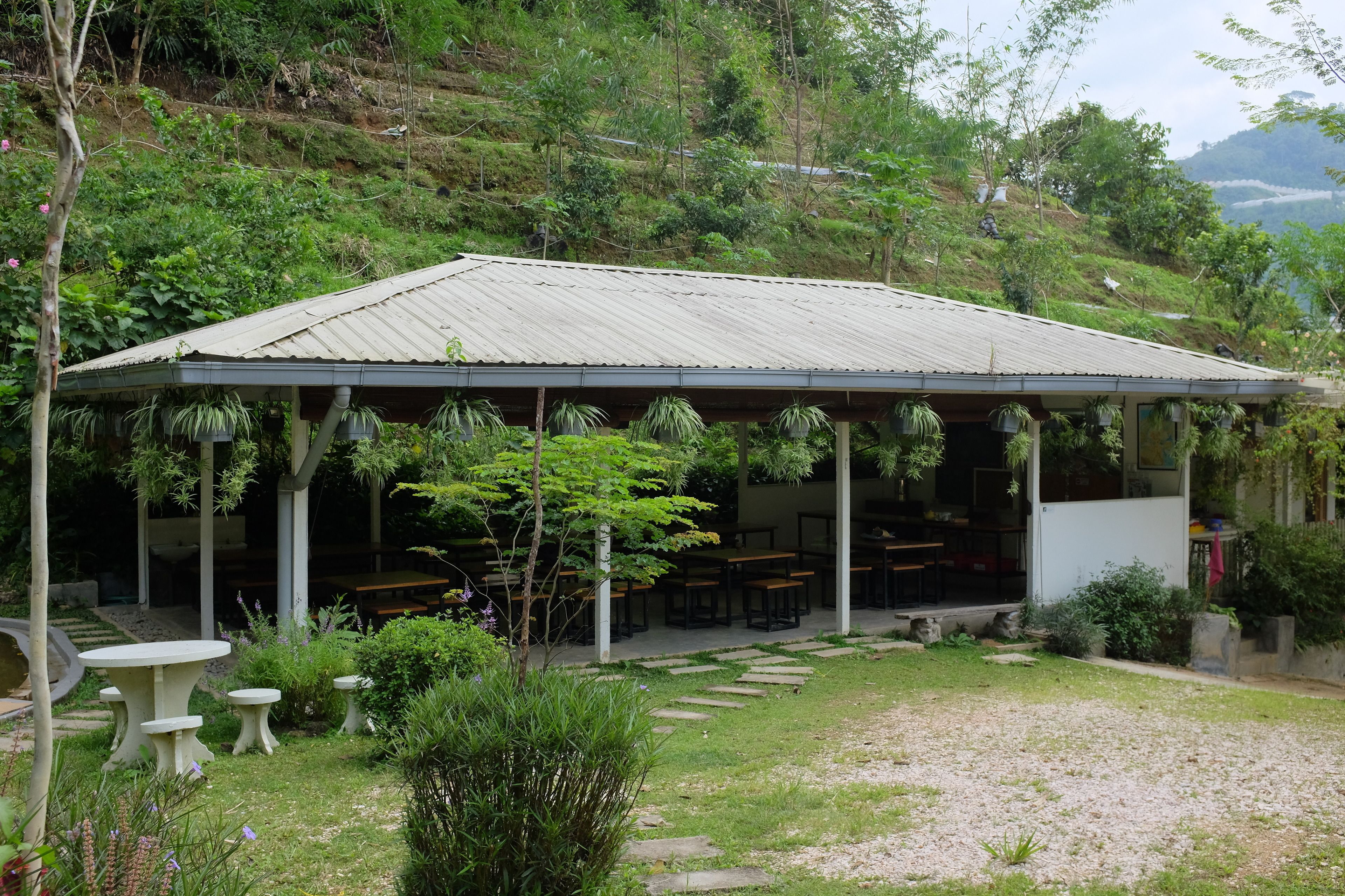 A covered outdoor seating area with tables and chairs, surrounded by lush greenery and a hillside backdrop.