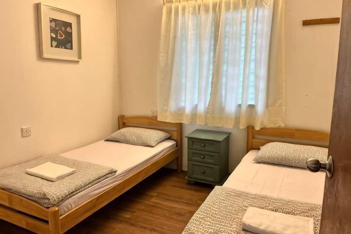 A room with two single beds, a small green nightstand, a framed picture on the wall, and a window with sheer curtains.