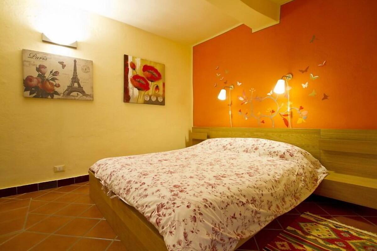 A bedroom with a bed, a nightstand, a wall-mounted lamp, and two wall art pieces.