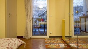 Una habitación con una cama, una alfombra estampada, y un ventanal corredizo que da a un espacio exterior con una mesa y sillas.