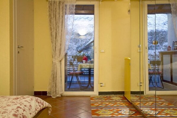 Una habitación con una cama, una alfombra estampada, y un ventanal corredizo que da a un espacio exterior con una mesa y sillas.