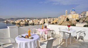 Una terraza con mesas y sillas blancas, con vistas a un pueblo costero con edificios y una masa de agua.