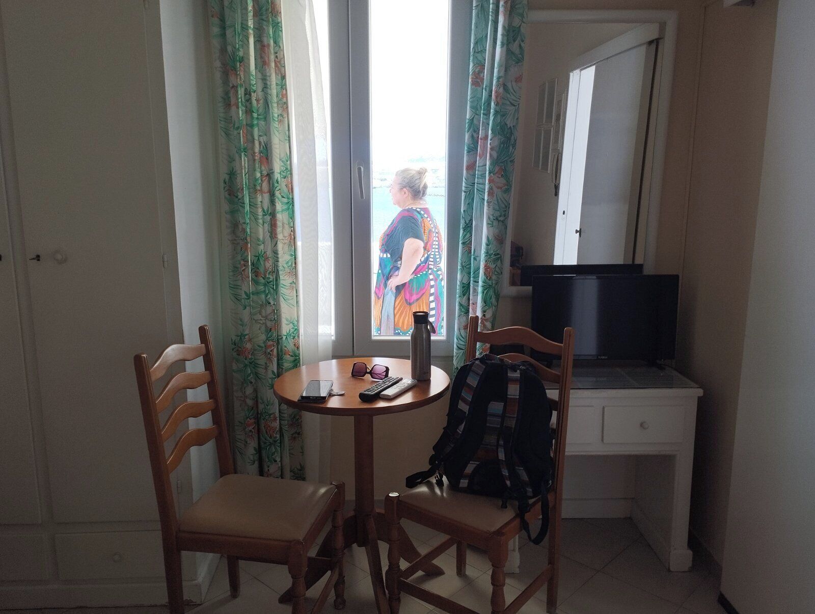 A room with a wooden chair, a small round table, a television, and a window with curtains.