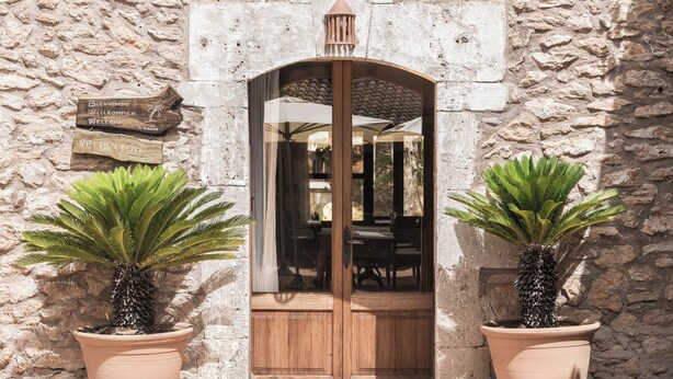 Une entrée en pierre, avec une porte en bois, deux plantes en pot et une pancarte de bienvenue.
