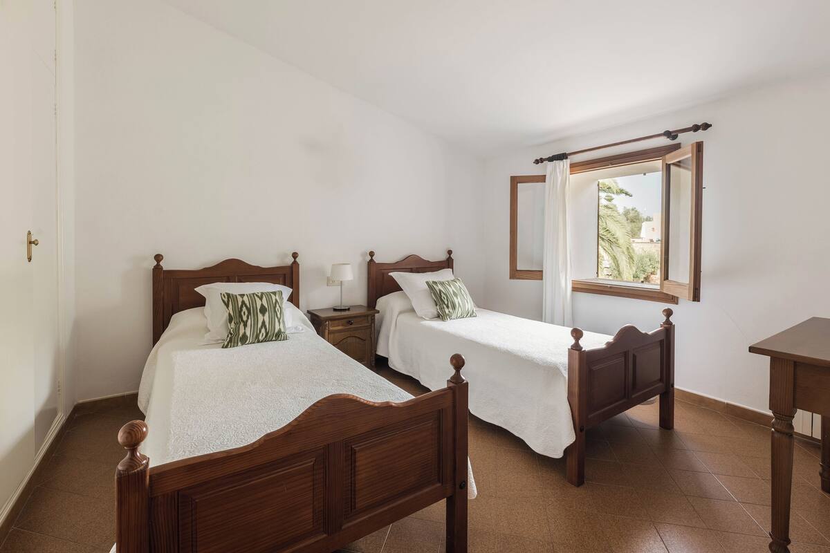 A bedroom with two wooden beds, a nightstand, a table, and a window with a view of greenery.