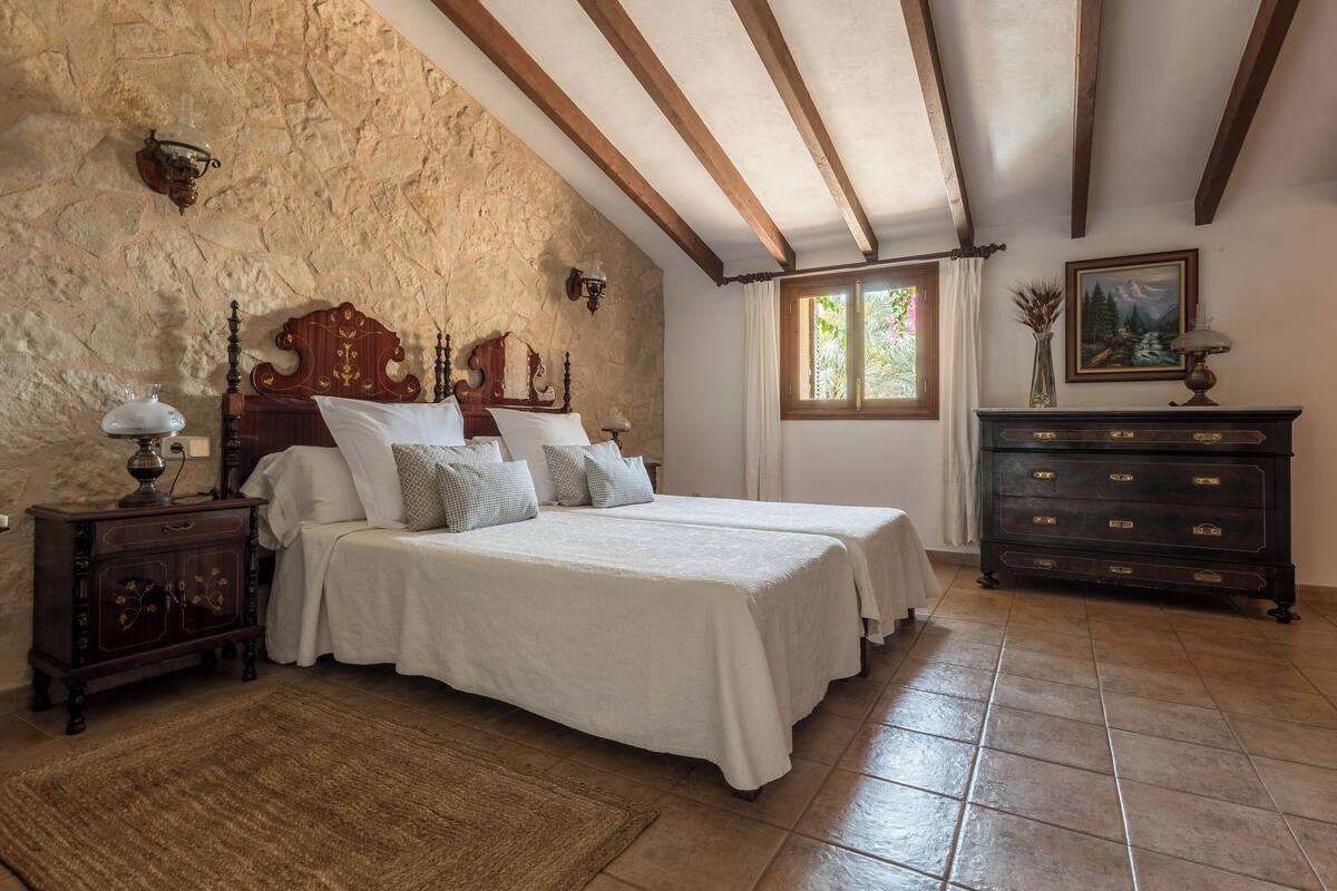 A bedroom with a stone wall, wooden beams, a bed with white linens, a nightstand, a dresser, and a window with a view of greenery.