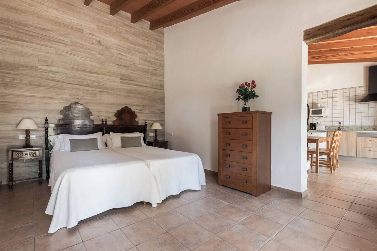 A bedroom with a large bed, wooden headboard, bedside tables with lamps, a wooden dresser, and a view into the kitchen area.