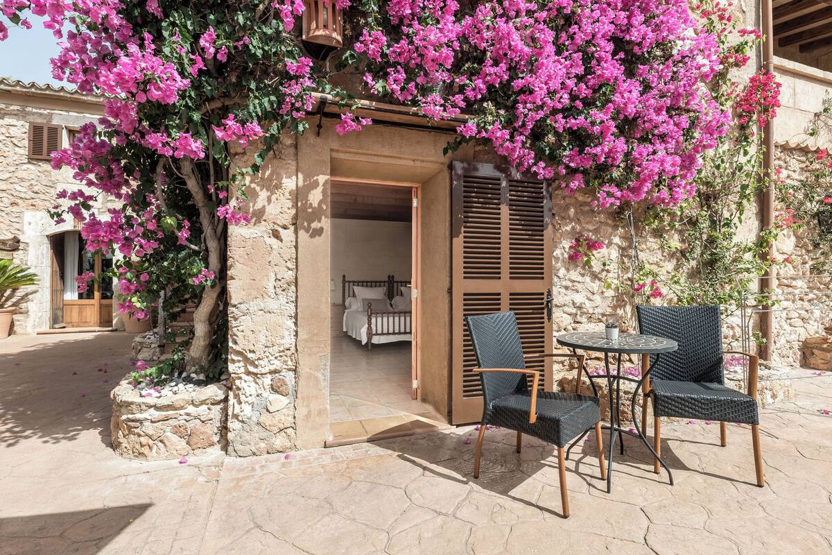 A stone building with a doorway leading to a bedroom. There are wicker chairs and a table outside.