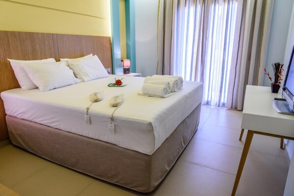 A neatly made bed with white linens and a brown headboard, a bedside table with a lamp, and a view of a window with curtains.
