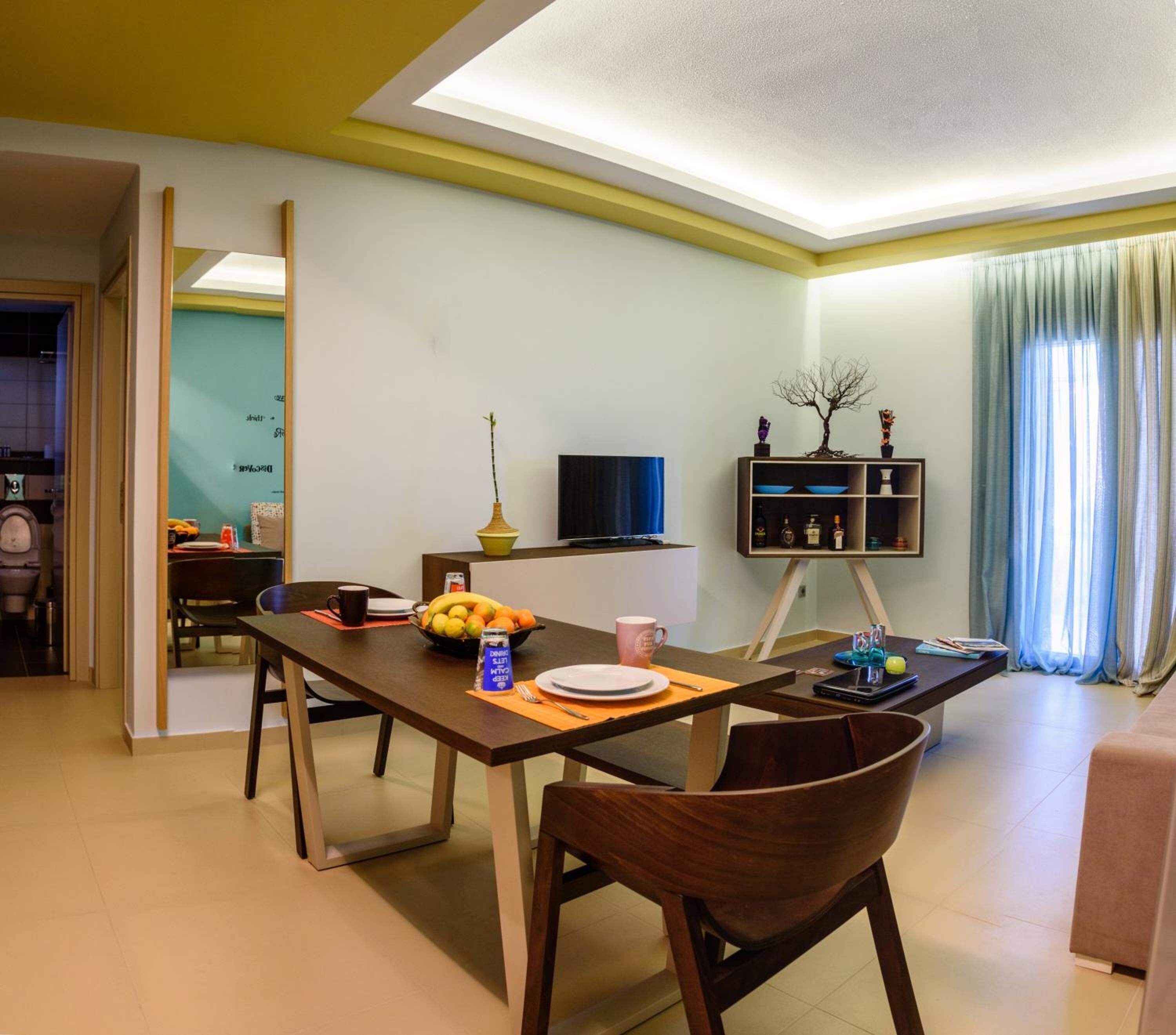 A modern living room with a dining area, a flat-screen TV, a bookshelf, and a window with curtains.