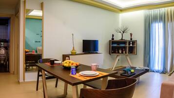 A modern living room with a dining area, a flat-screen TV, a bookshelf, and a window with curtains.