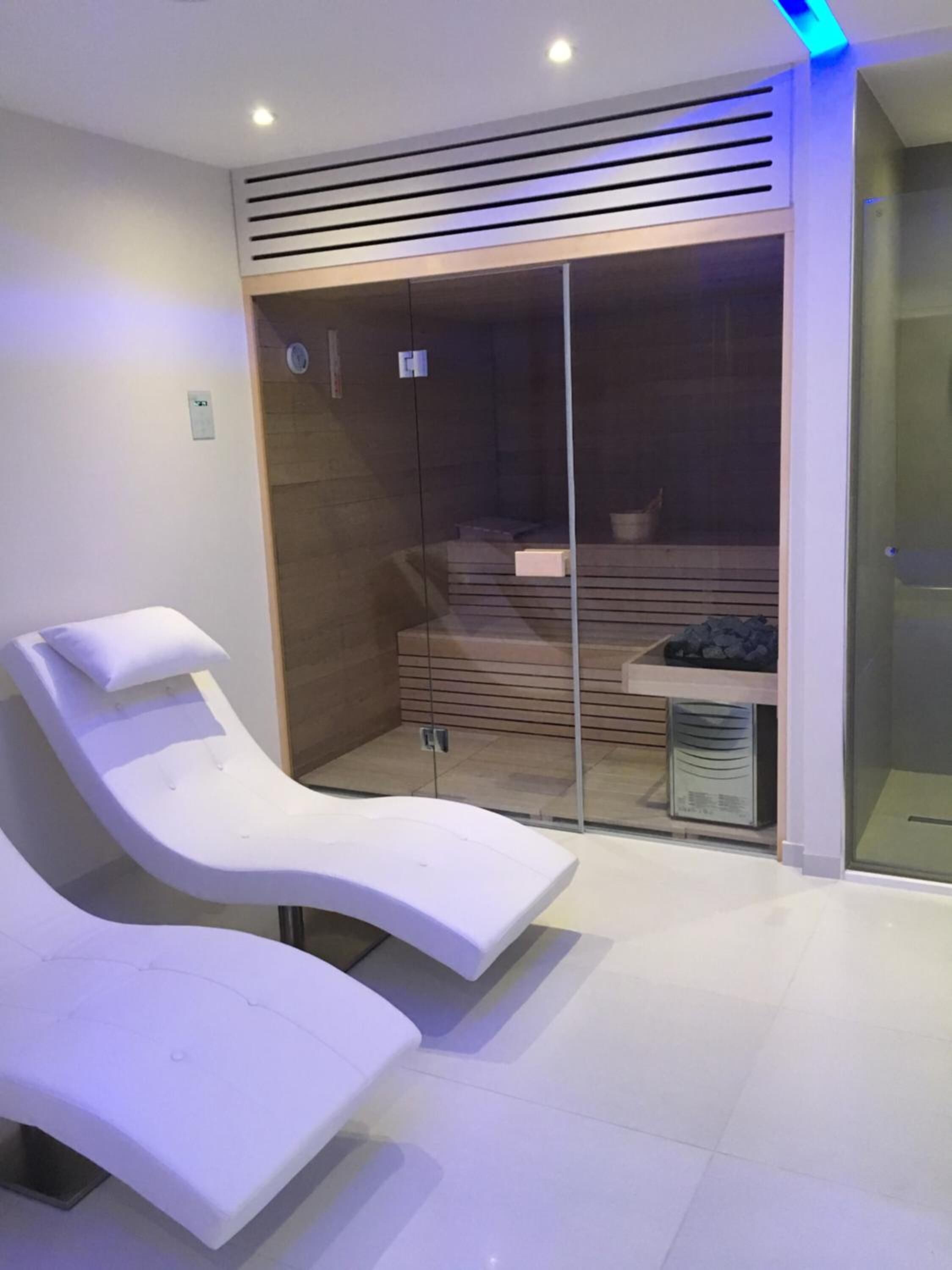 A modern spa room with two reclining chairs, a glass-enclosed shower area, and a wooden slatted wall.
